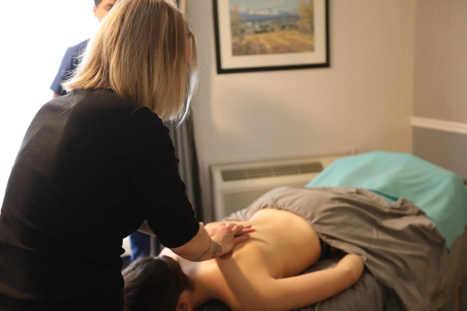 Deidra giving a back massage in a bright treatment room with a framed painting on the wall