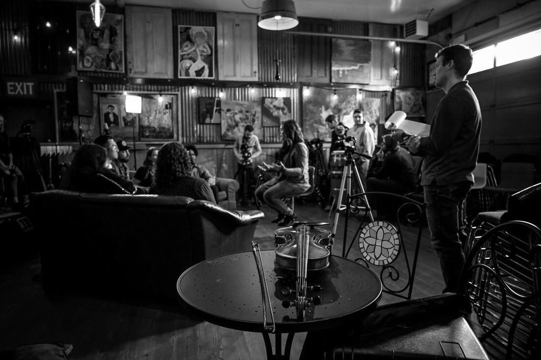 Black and white photo of a speaker reading to a small audience in a café, with a camera on a tripod