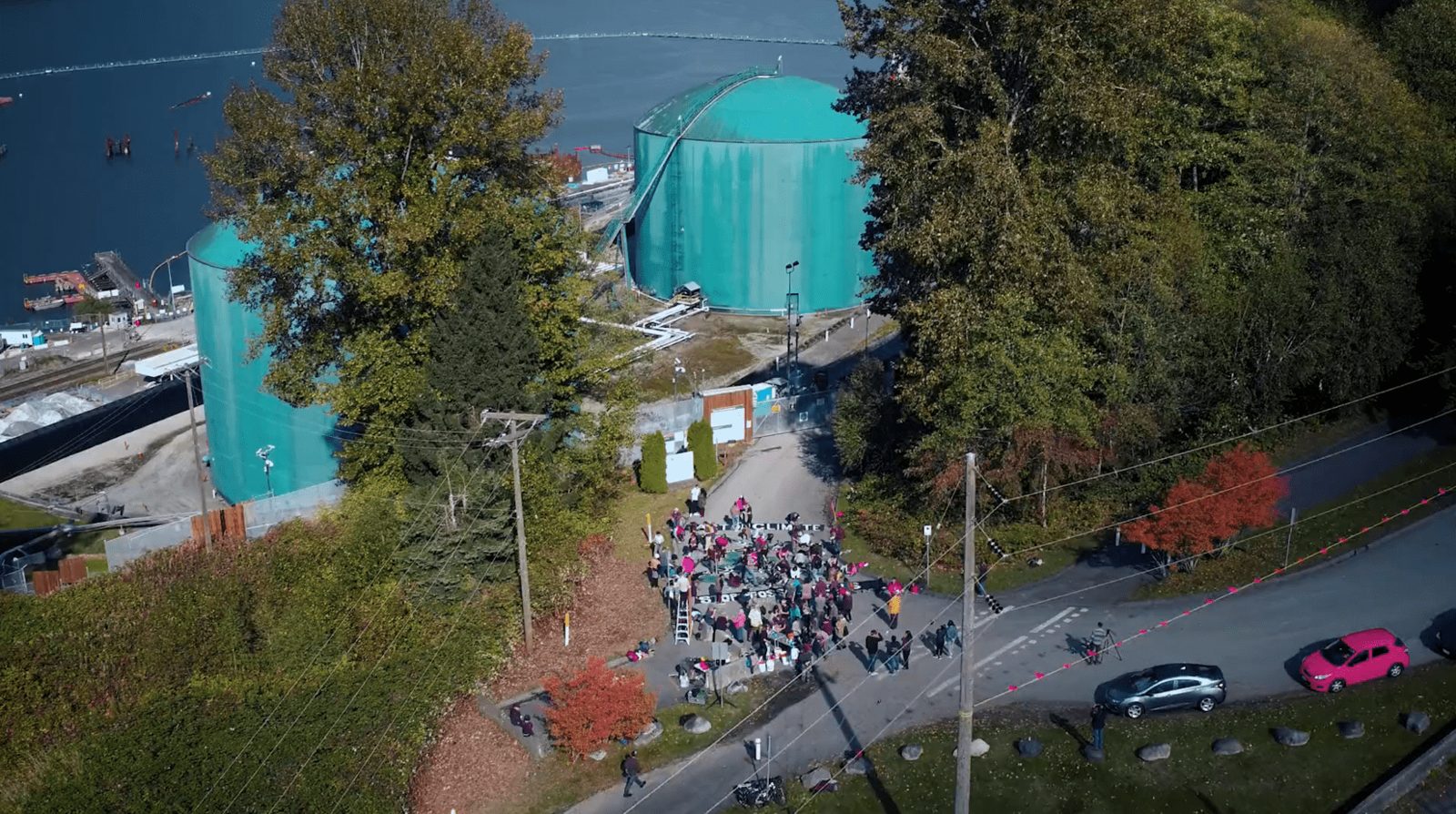 A crowd gathered on a road below teal fuel storage tanks, filmed by drone, from the Salish Sea art action