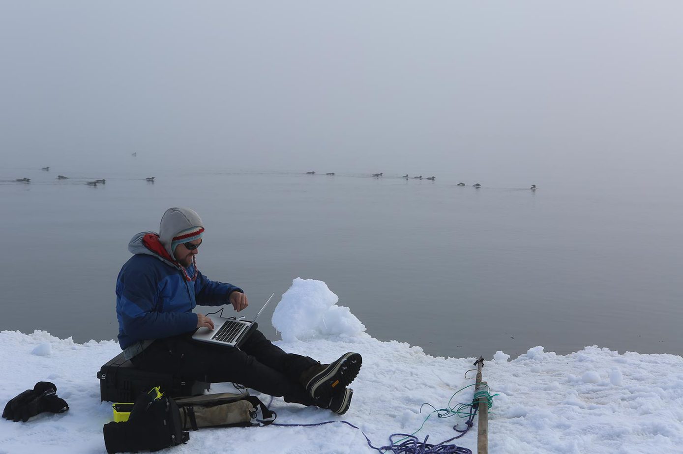 A man in winter gear working on a laptop at the edge of the sea ice