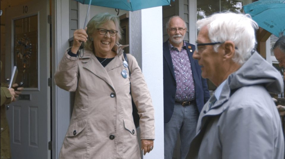 Three people smiling and talking on a doorstep under teal umbrellas, from the George Orr campaign video