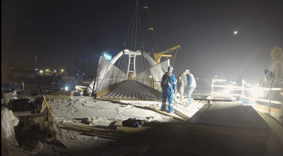 Workers in blue coveralls beside a lit bridge span at night, from the Salton Bridge timelapse